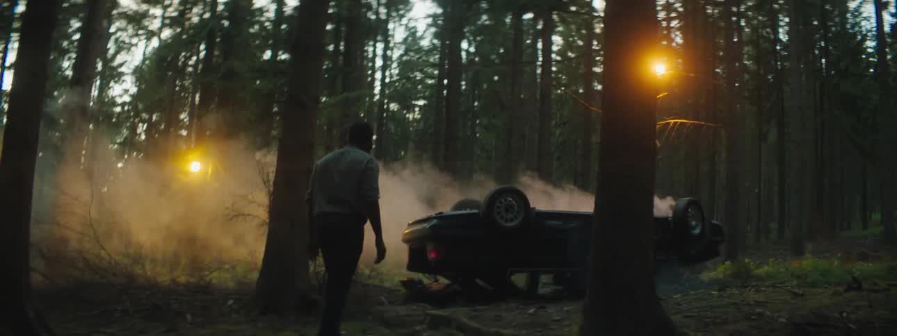 A man walks through a forest at dusk towards a flipped car that appears to be smoking and badly damaged