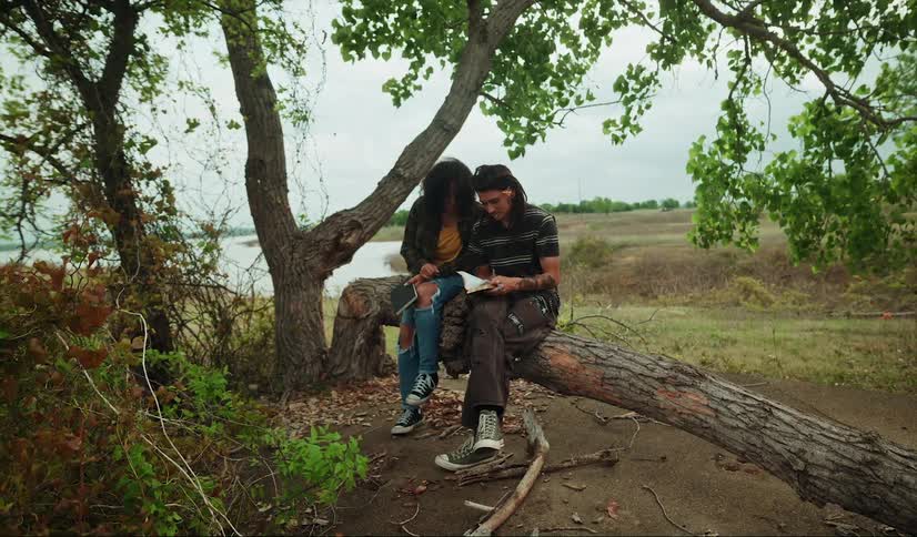 A man and woman are sitting on a tree trunk near a river looking in a book