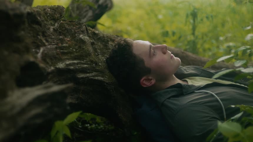 Young rosy cheeked man rests against moss covered rocks in the woods