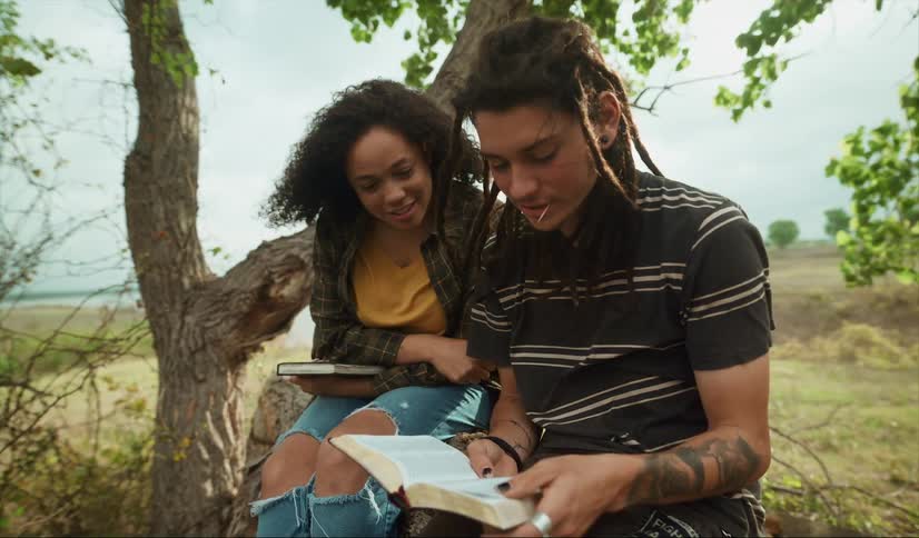 Man and woman sit outdoors near a tree talking over a book with water in the background