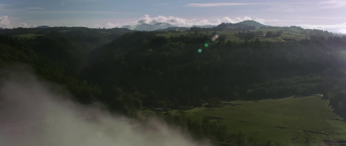 Low lying clouds float over field and water with forest and hills stretching to the horizon