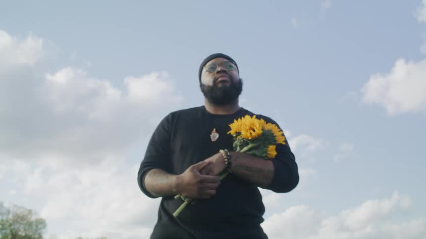 A man walks outside carrying a bouquet of flowers and solemnly looks down at the ground