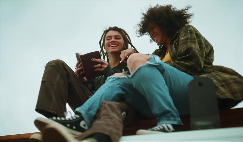 Man with dreadlocks rings brown pants holding a brown notebook sits next to woman with curly hair plaid shirt and ripped jeans laughing