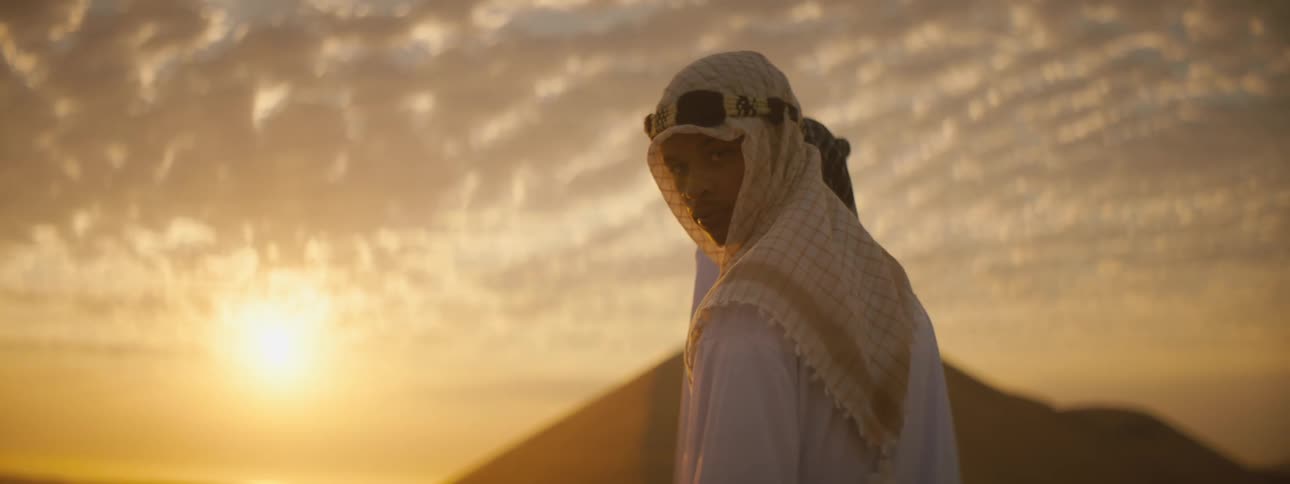 Man wearing head covering that flutters in breeze stand back lit by sun turning sky red