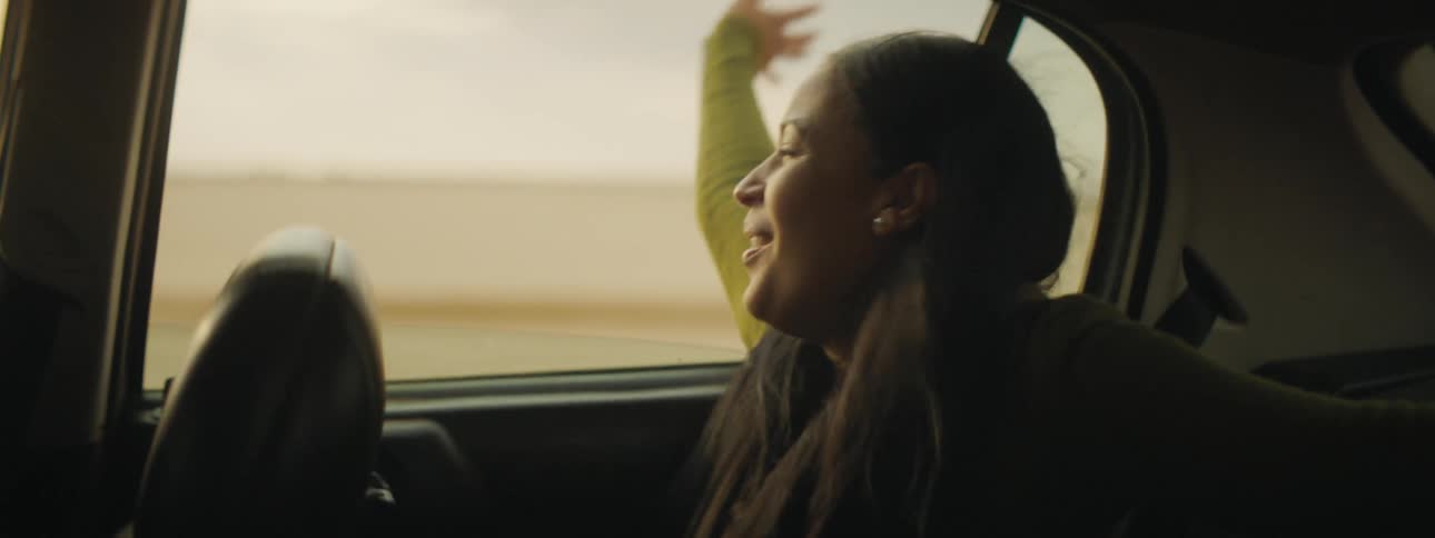 A woman in the backseat of a car laughs as she holds her arm out the window as she goes by fields
