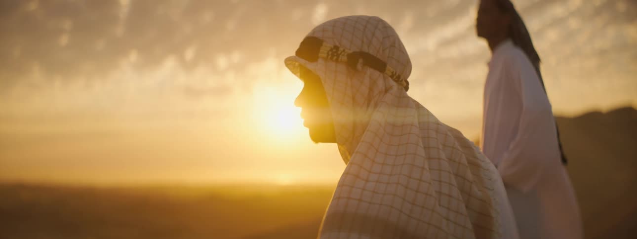Two men look into the distance on the desert while the sun sets behind them with mountains in the distance