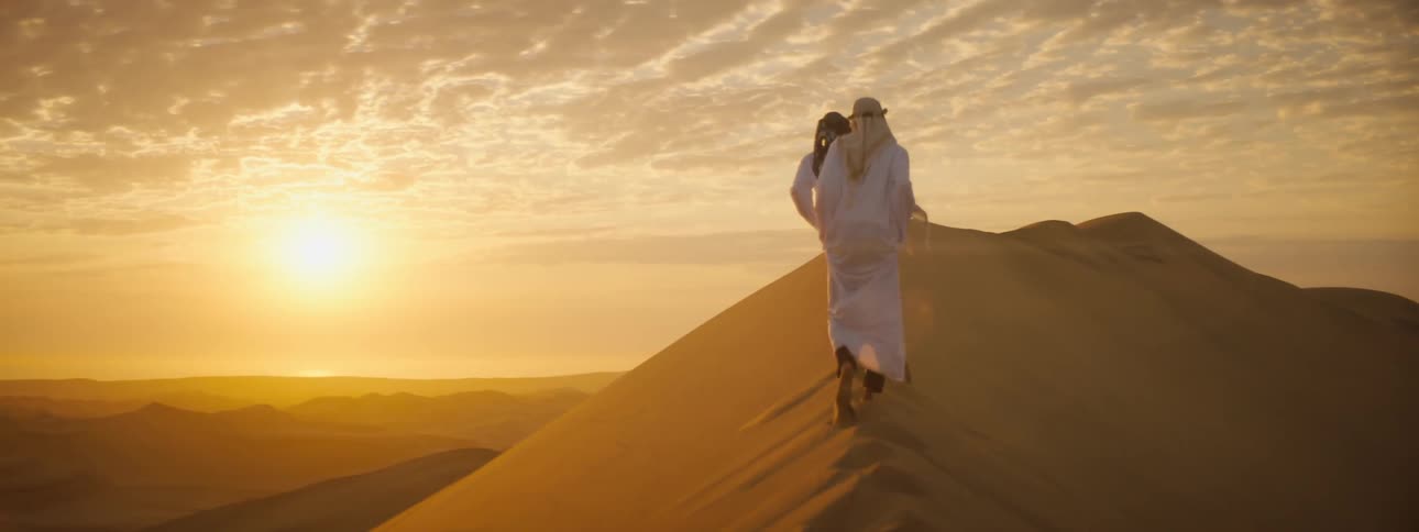 Two people in robes and turbans walk up a sandy hill in a desert as the sun sets