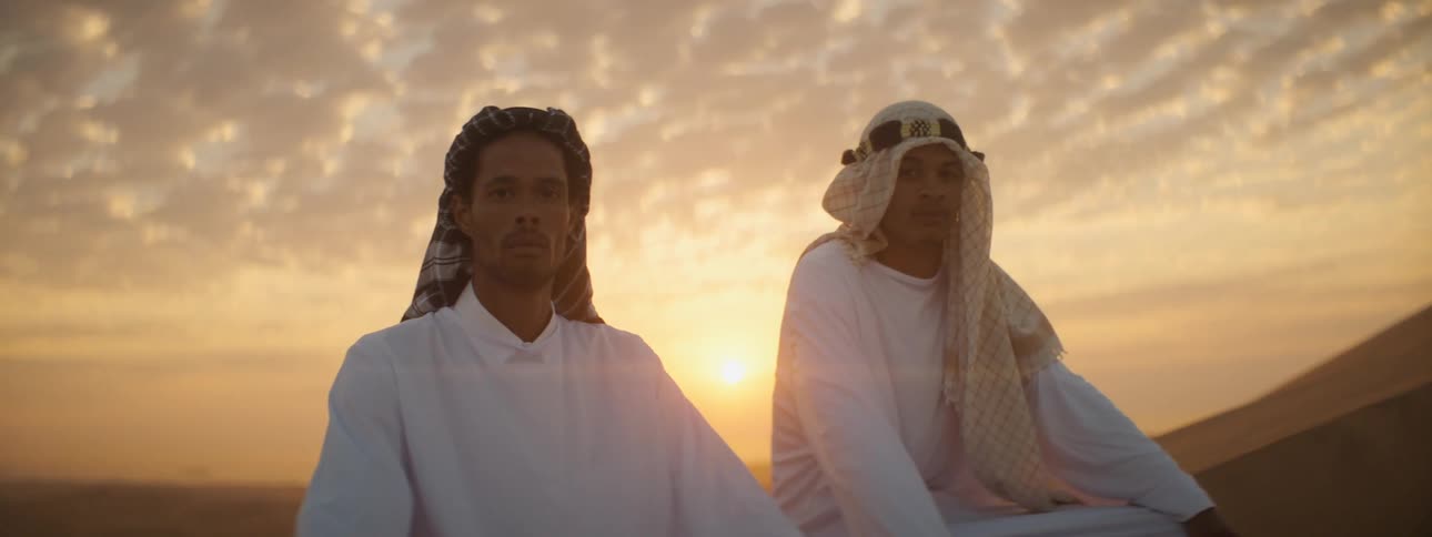 Two men wearing white and headdresses sit on sand dune in desert as sun turns sky red