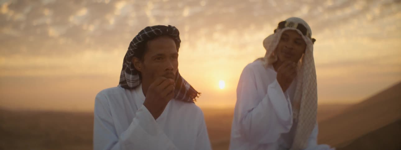 Two middle eastern men in robes sitting in the desert and eating from their hands
