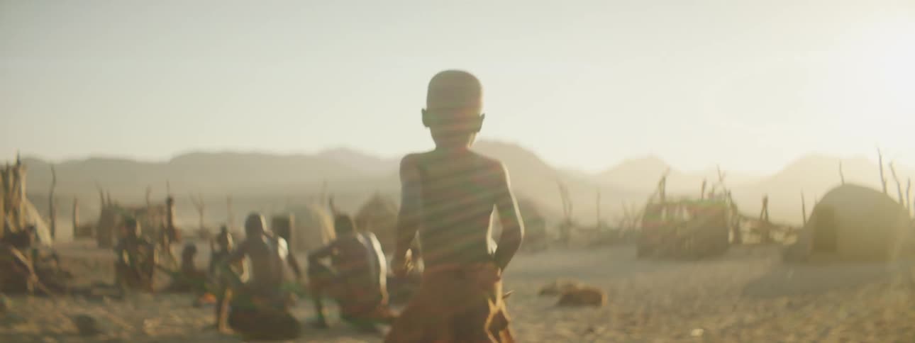 A young boy jogs out of a small hut in his village and joins others gathered outside