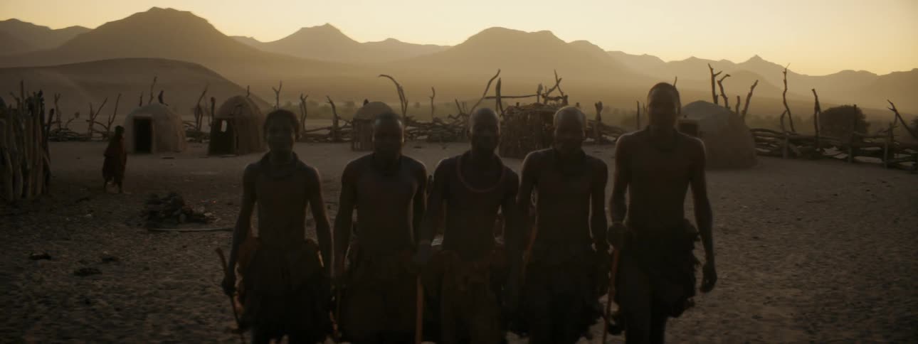 Men wearing traditional attire walk forward in a group in village of huts with misty mountains on horizon under tinted sky