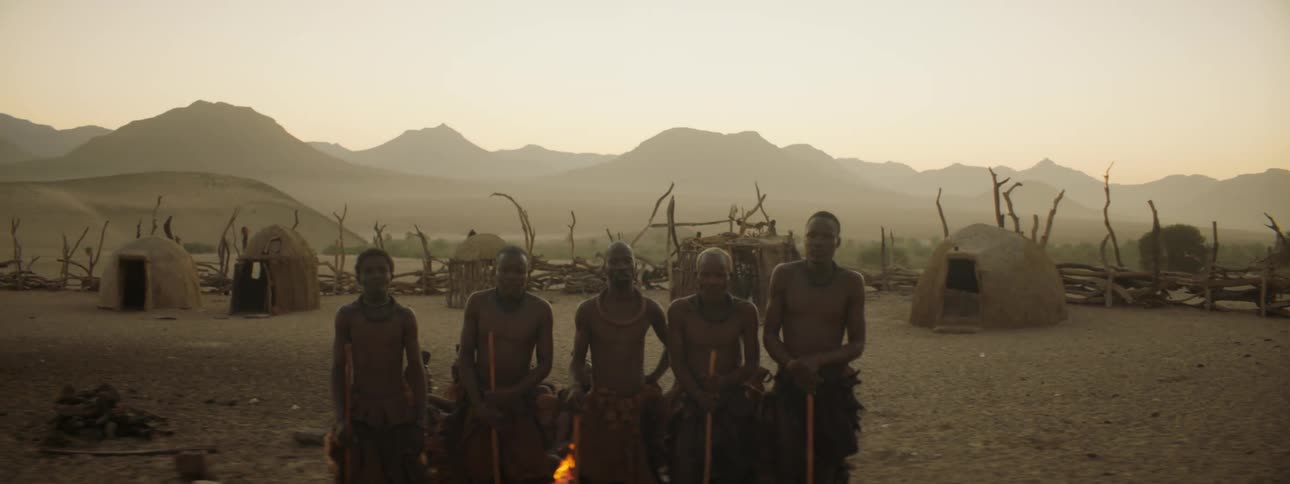 Five people sit behind a small fire behind them are small huts with large branches and rough fencing around them behind that is a mountain range where you can see the sunrise
