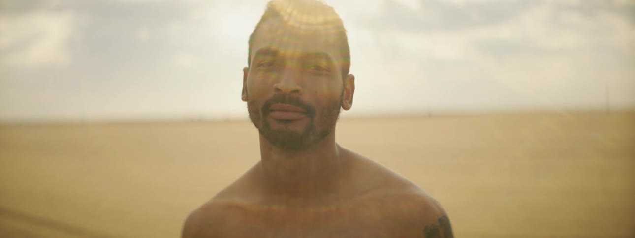 Man with a shoulder tattoo and a beard stands on the sand and looks at the camera on the beach