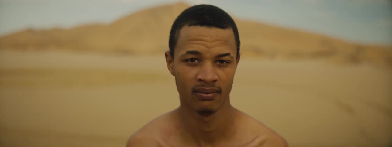 Man stands bare chested on sand stretching toward hill under light blue sky