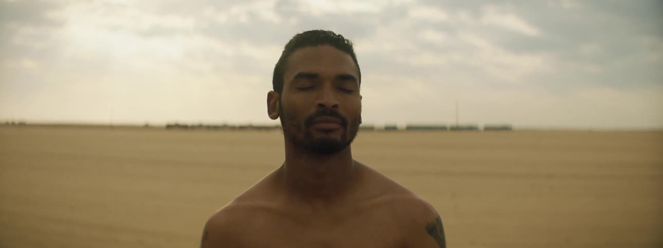 Man with a shoulder tattoo and a beard stares at the camera while a train travels in the background