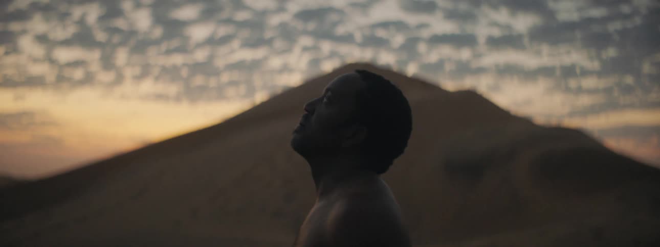 A man standing outside with a bare chest in front of a hill looking up at the sky