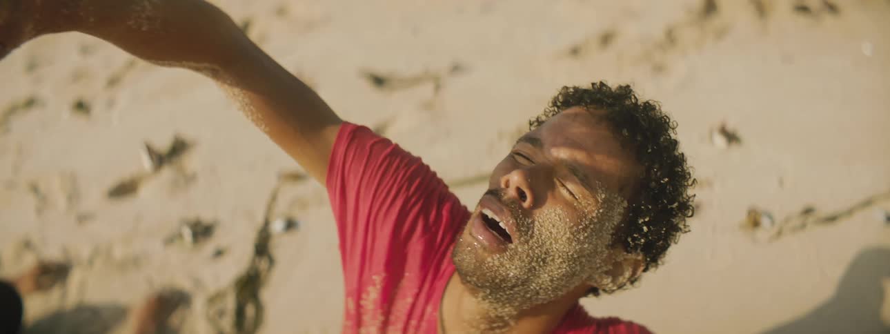 One man sitting in the desert holds his hand up to block his eyes from the sun as he looks up at the sky as another man lies face down in the sand