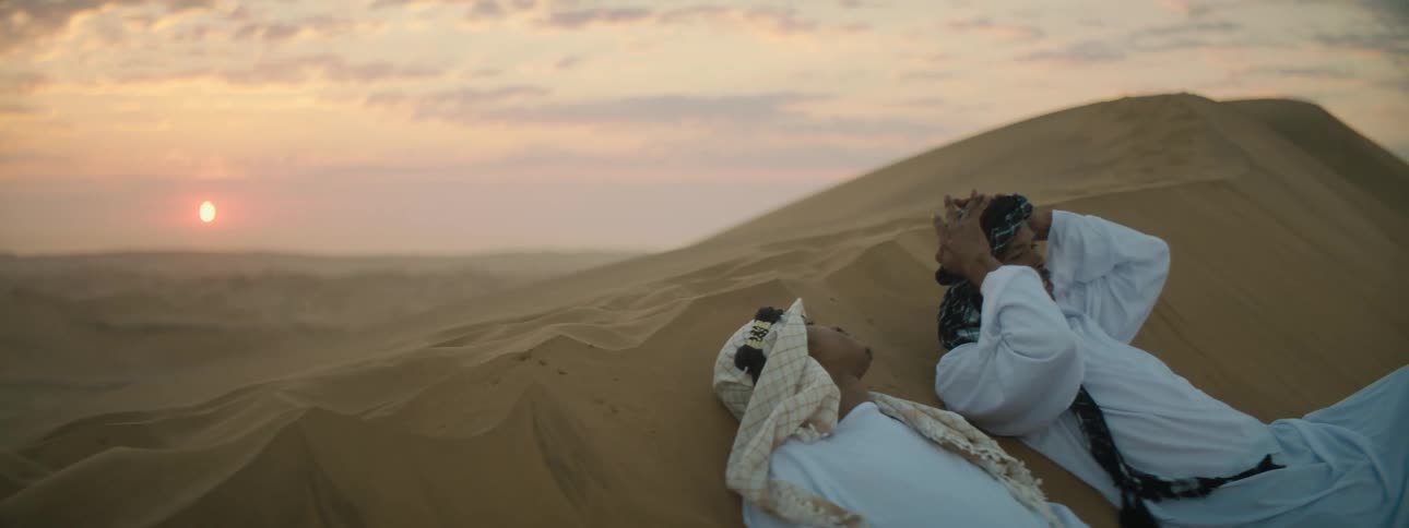 Two men are relaxing on a sand dune pointing at the sky with the sun low in the sky