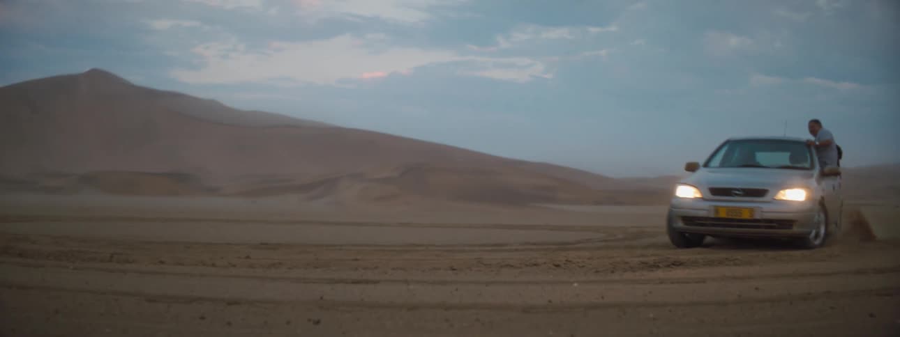 Two people hand out the windows of a vehicle as it does donuts in a desert terrain
