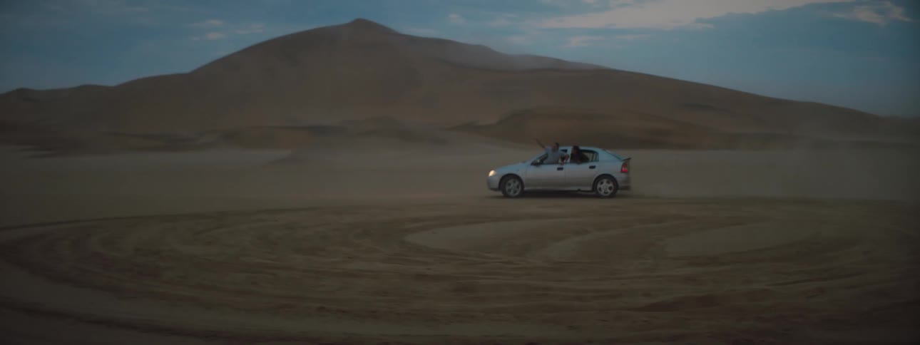 Two people hang out of car windows while the vehicle is doing donuts in a desert terrain area
