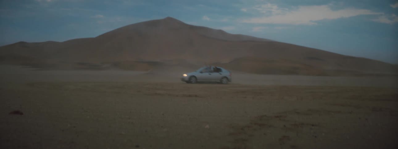 A person hanging out the window of a vehicle driving in the desert with the sky and clouds in the background