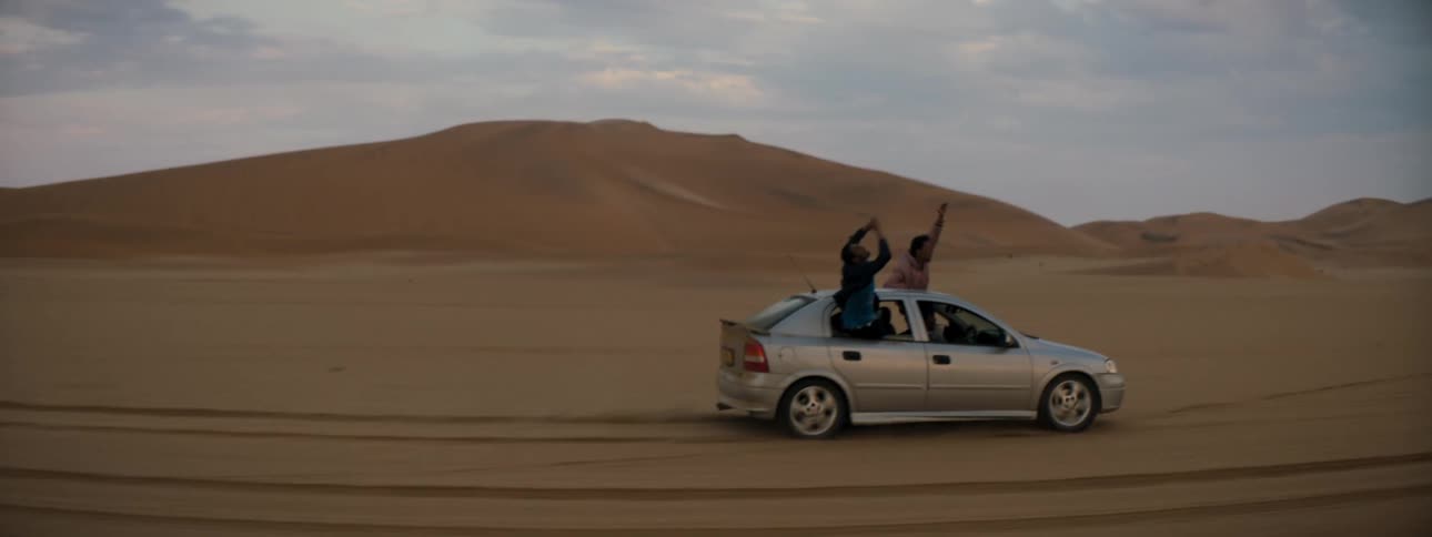 People hanging out of a car window and shouting as it drives across the desert