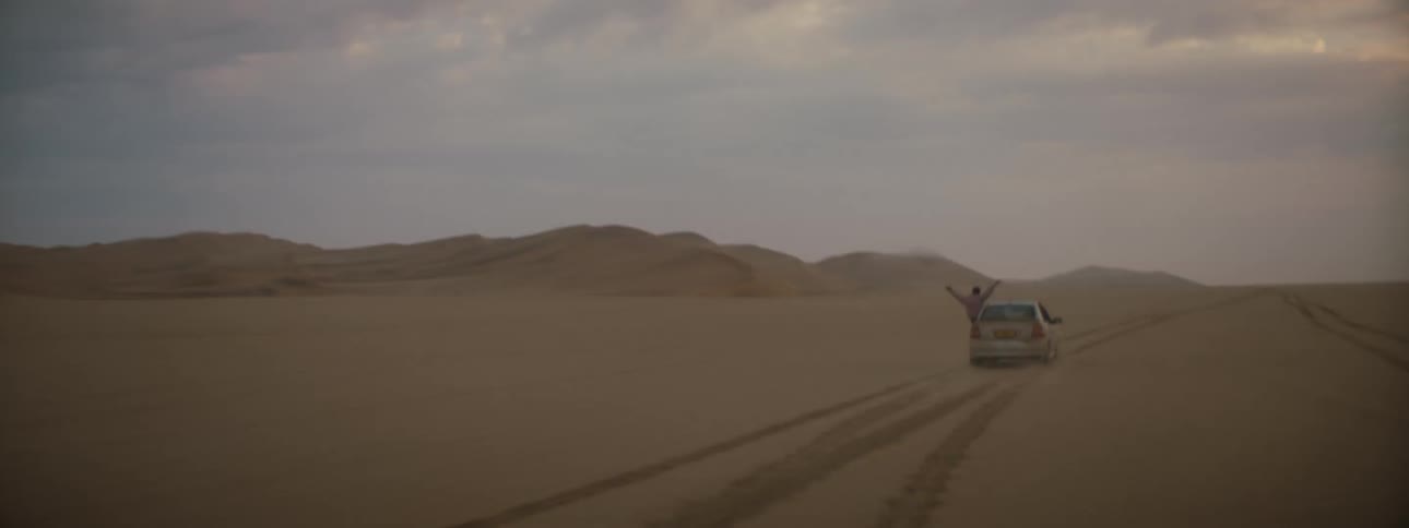 A person hanging out the window of a vehicle driving in the desert with the sky and clouds in the background