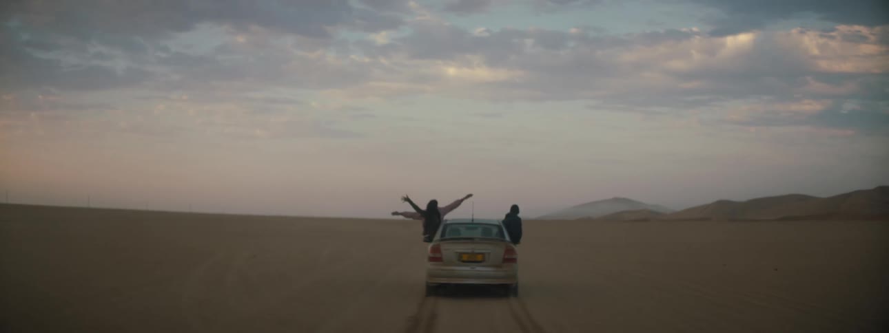 A vehicle driving in the desert with people hanging out the window with the sky and clouds in the background
