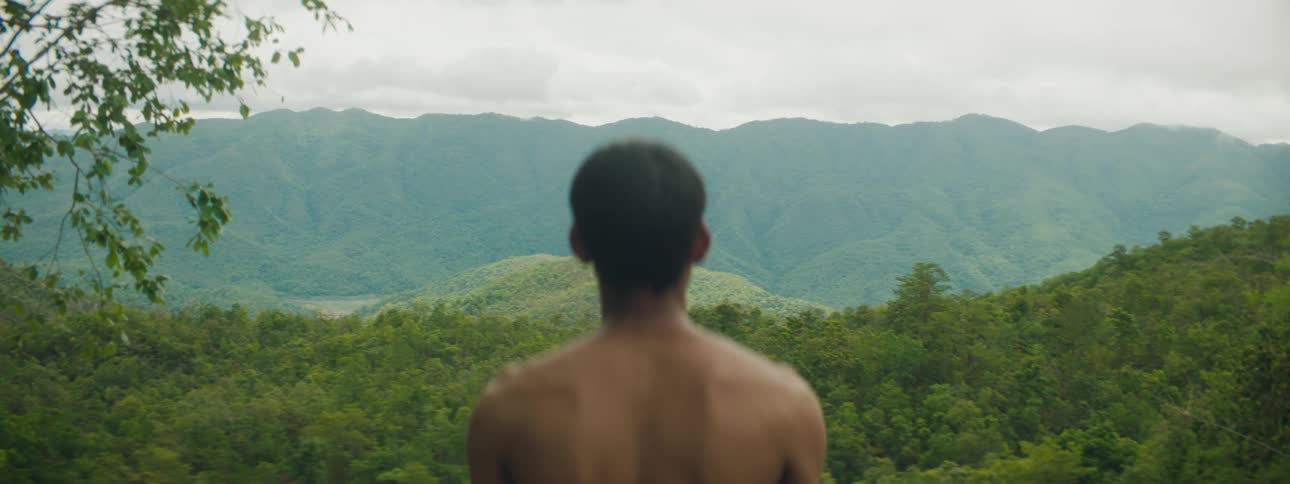 A man stands shirtless outside and looks at bush covered hills