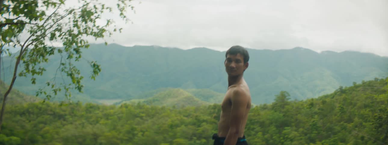 A shirtless young man runs to the edge of a cliff and gaze out over the forested countryside before turning to gaze into the camera