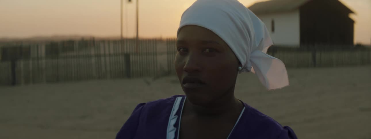 A woman in a blue dress and a white headwrap stares into the camera while standing outdoors in a rural area in the evening