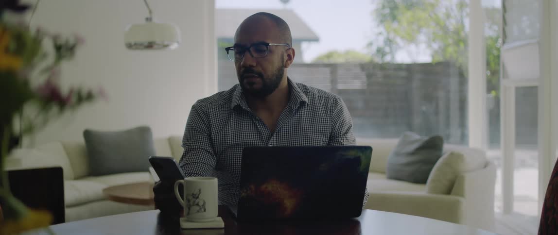 A man sits at home and uses his laptop and then picks up his cell phone and looks at it