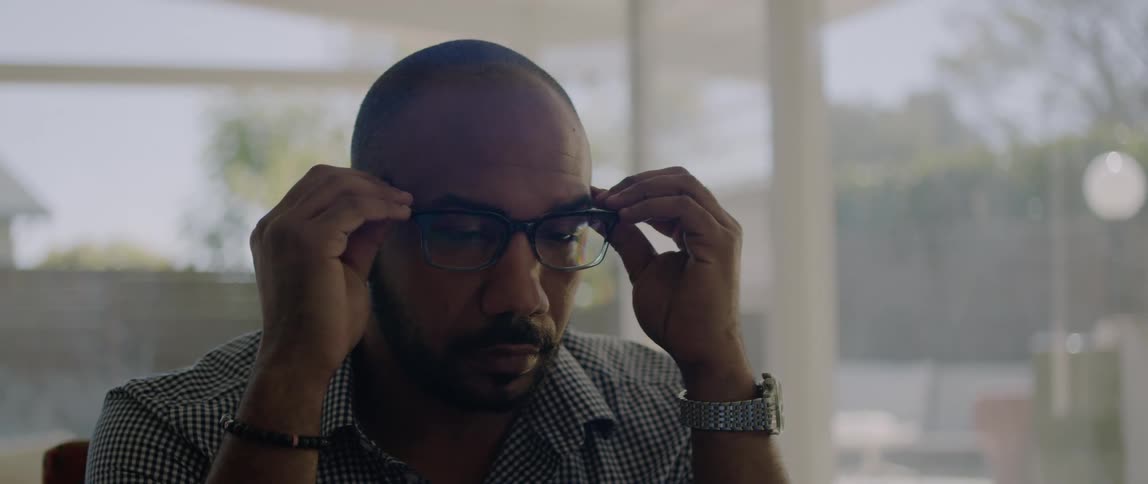 A man slowly puts his glasses back on while hes seated indoors