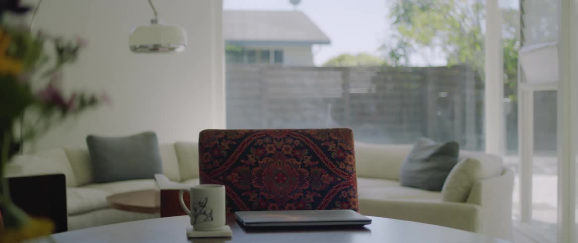 There is a laptop computer and a coffee cup on a table in front of a chair and a couch and a window behind it