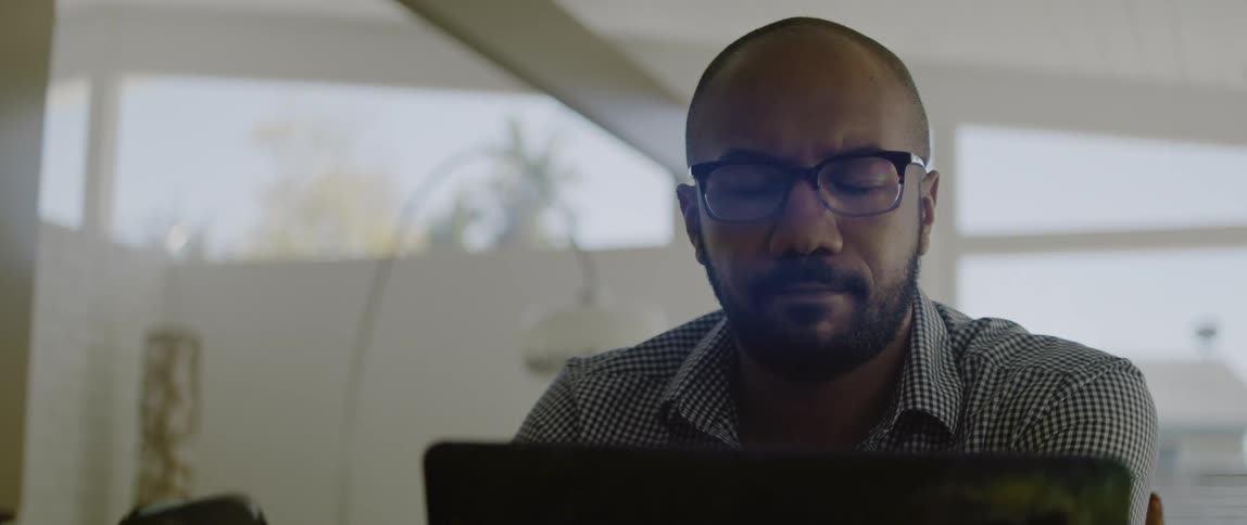 A man wearing glasses looks down at his laptop while holding his phone