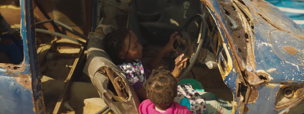 Black girl sits in an old rusty vehicle and pretends to drive while a black toddler stands next to her