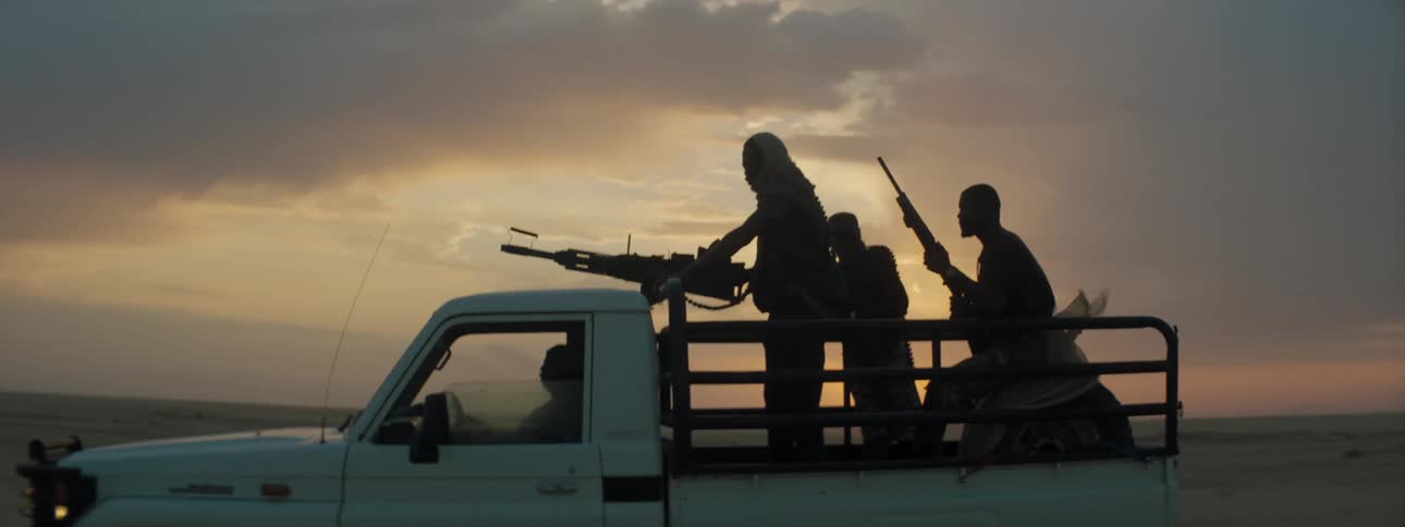 Men ride in truck bed holding weapons as sun turns sky on horizon red