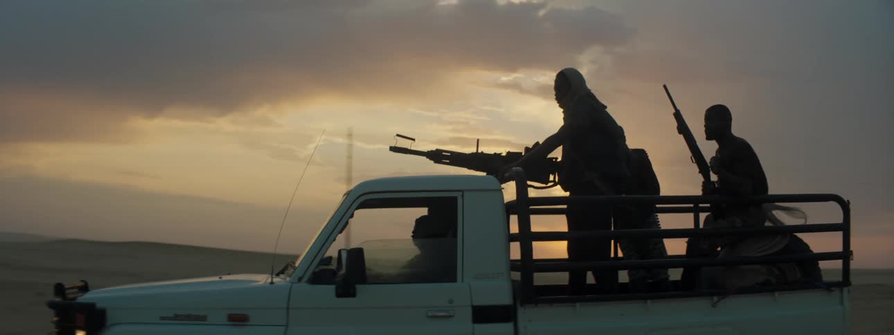 A group of armed men travel in the rear of a truck of as it travels down a road during the evening