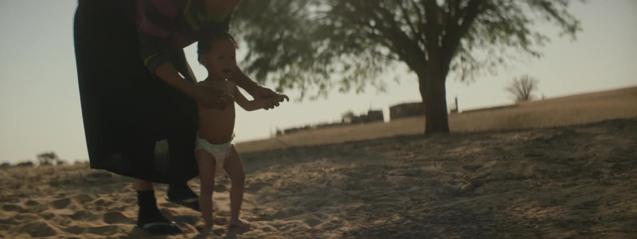An elderly woman helps a baby walk through the sand in front of a field