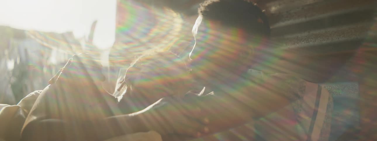 A man and woman lie in bed in a shelter with no wall and he kisses her head as she rubs his chest