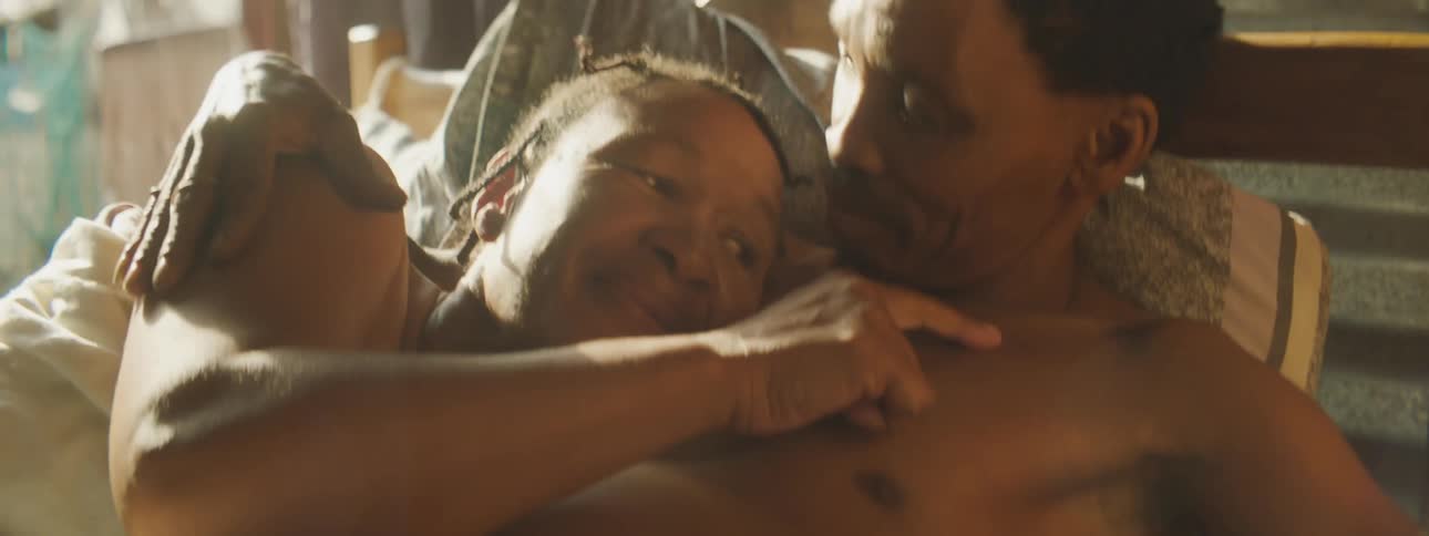 A black man and woman snuggle together on a bed in a dimly lit room