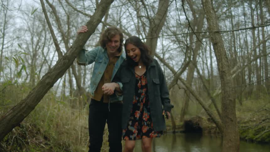 A teenage couple hold hands while walking along a creek in the woods