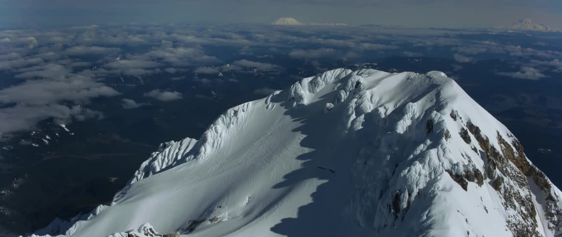 The sun shines on a snow covered mountain peak high above the clouds and there are snow covered mountains in the distance