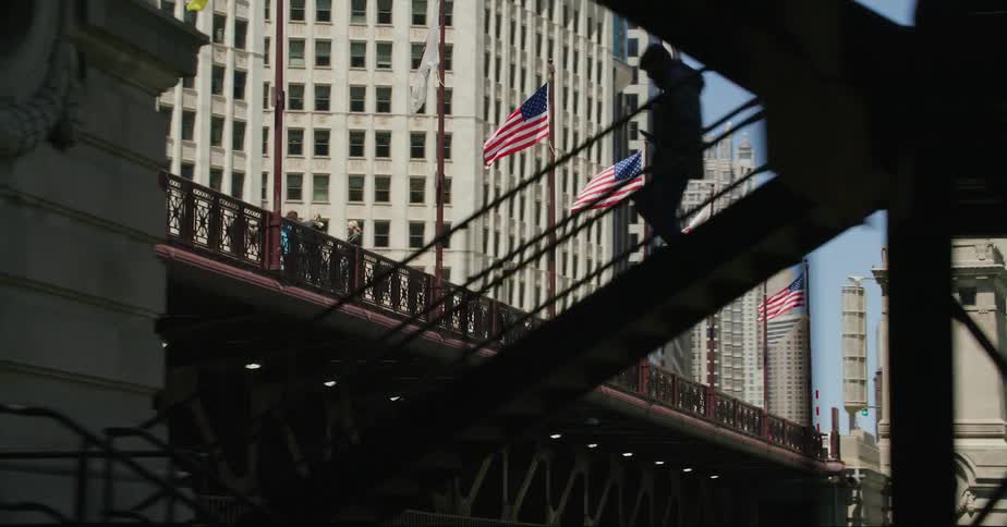 A row of flags wave on poles placed along a bridge in a metropolis during the day