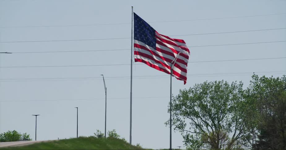 An american flag waves in the wind on a pole in a park among trees as traffic passes on the road