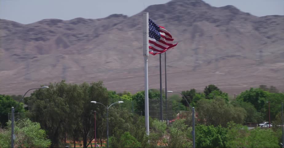 An american flag waves in the wind on a pole on the roadside as traffic passes by and there are mountains in the distance