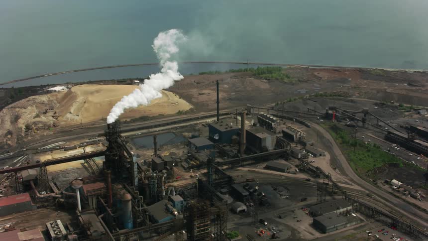 Smoke exits chimney at plant sitting on shore next to to mound of sand