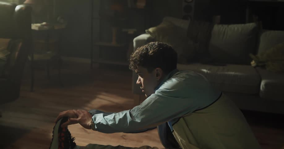 Man with curly hair light blue jacketbrown pants and sneakers sits on floor stretching his legs in living room at home