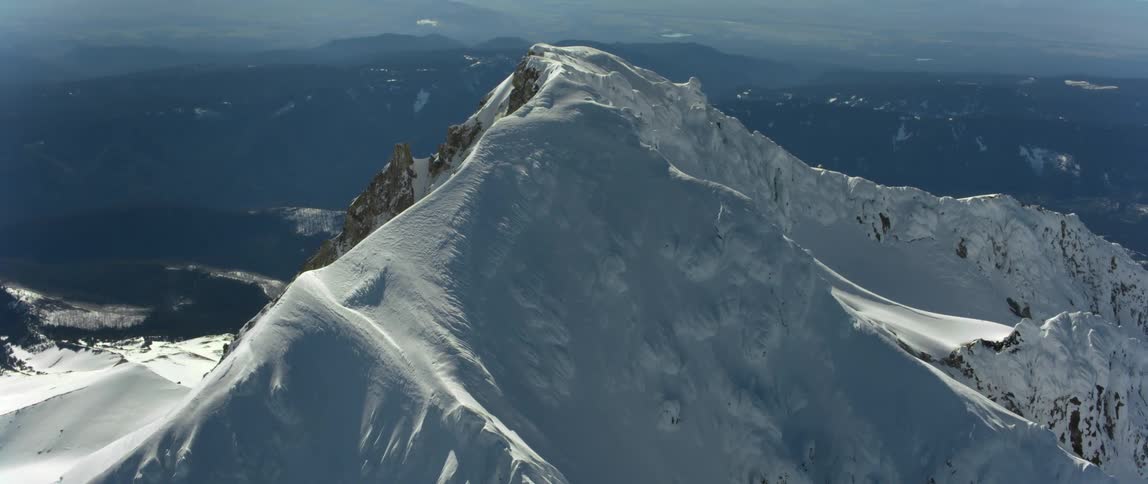 The sun shines on a snow covered mountain high above other mountains in the distance