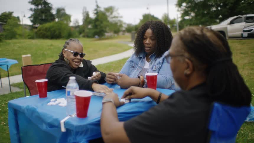 Three women are sitting outside at a card table playing a scrabble game